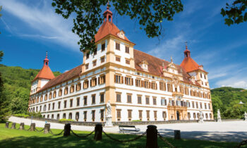 Ein großer, reich verzierter Barockpalast mit roten Dächern und weißen Wänden steht unter blauem Himmel, umgeben von Grün und Statuen.