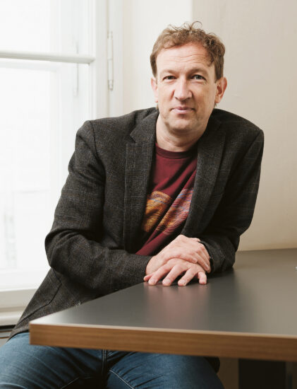 Ein Mann in Blazer und Jeans sitzt an einem Schreibtisch am Fenster, auf dem Tisch stehen ein Glas und ein Notizblock.