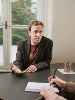 Ein Mann in einer Jacke gestikuliert, während er an einem Schreibtisch spricht, während eine andere Person mit Stift und Notizbuch Notizen macht. Im Hintergrund sind ein Fenster und Bäume zu sehen.