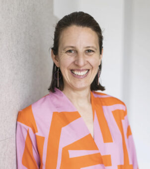 Eine lächelnde Frau mit braunen Haaren in einer rosa und orange gemusterten Bluse und einer schwarzen Hose steht mit einer Hand in ihrer Tasche vor einem hellen Hintergrund.