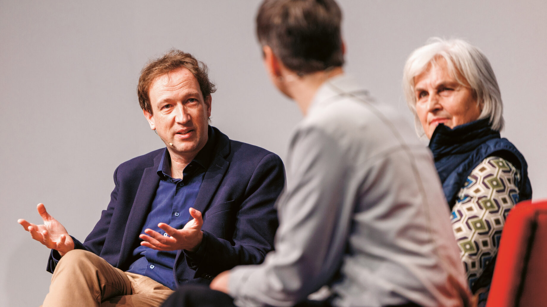 Drei Personen sitzen bei einer Podiumsdiskussion und sprechen. Ein Mann gestikuliert beim Sprechen, die anderen hören aufmerksam zu. Alle scheinen in ein Gespräch vertieft zu sein.
