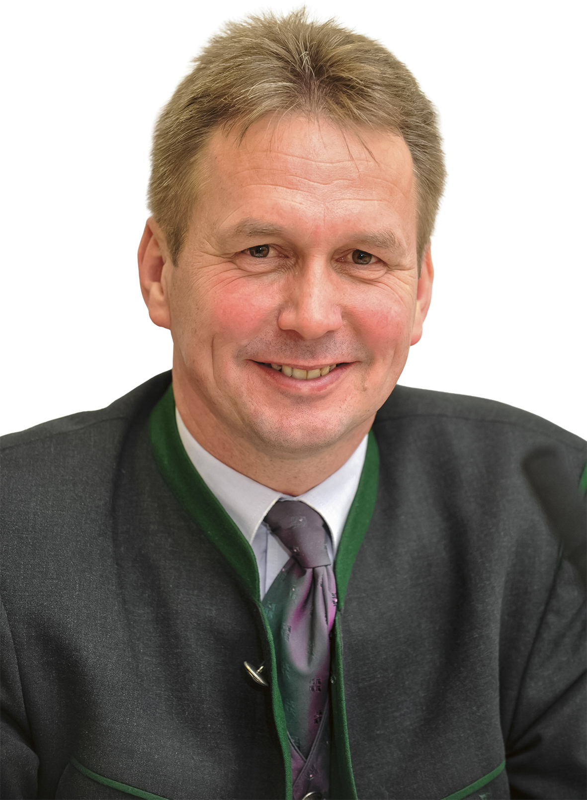 Lächelnder Mann in dunkler Trachtenjacke mit grünem Saum und Krawatte, vor weißem Hintergrund.