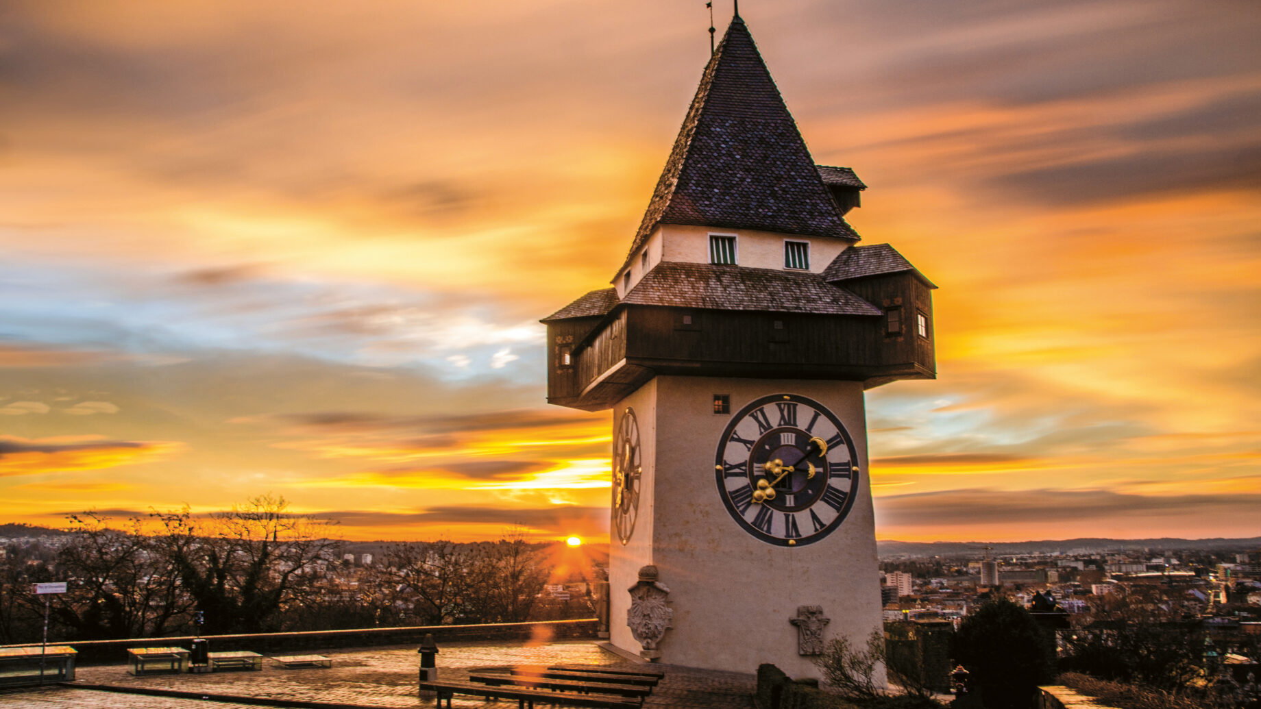 Ein historischer Uhrenturm steht auf einem Hügel bei Sonnenuntergang, während dramatische Wolken und goldenes Licht den Himmel und die Stadt darunter beleuchten.