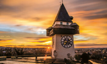 Ein historischer Uhrenturm steht auf einem Hügel bei Sonnenuntergang, während dramatische Wolken und goldenes Licht den Himmel und die Stadt darunter beleuchten.