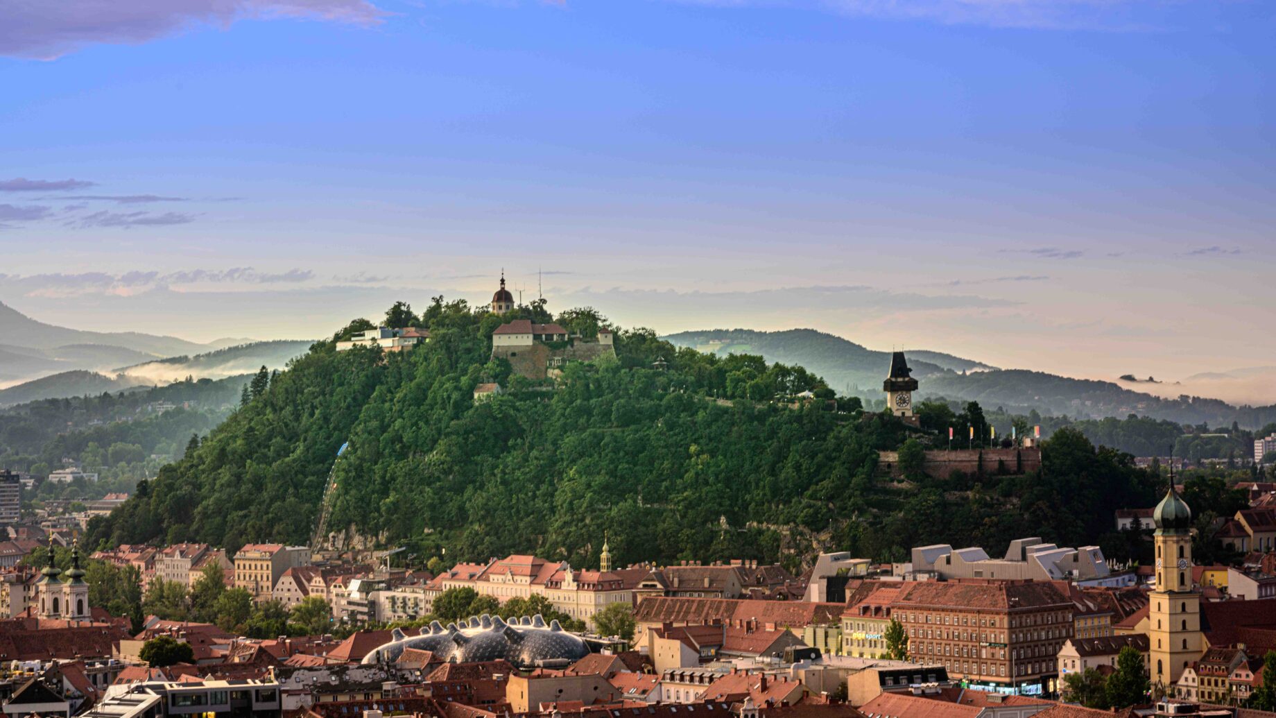 Ein üppig grüner Hügel mit historischen Gebäuden erhebt sich über Graz, wo die Battery Innovation Days unter einem farbenfrohen Sonnenaufgangshimmel die Zukunft der Steiermark beflügeln.