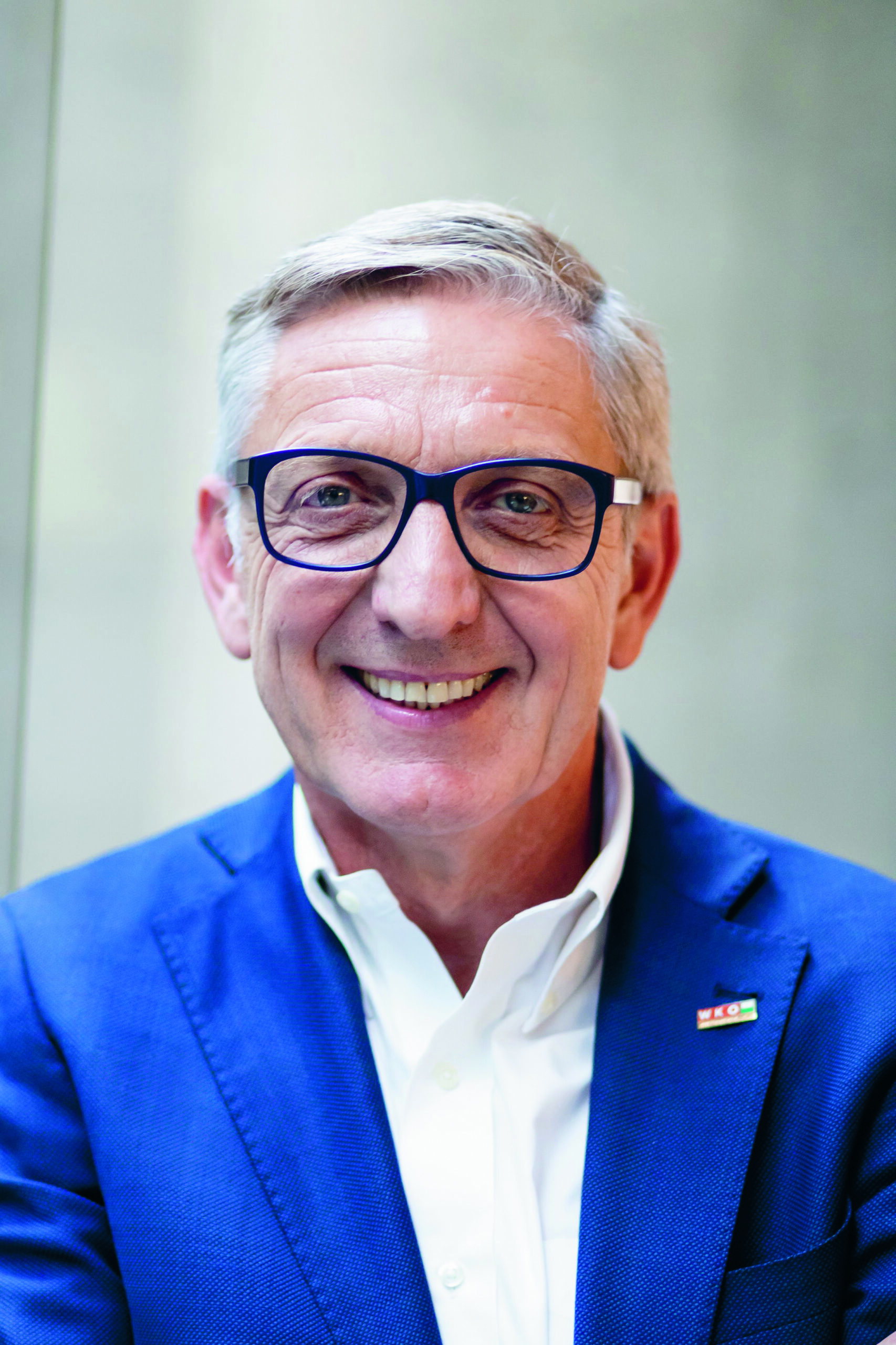 Lächelnder älterer Mann mit grauem Haar, Brille und einer blauen Anzugsjacke über einem weißen Hemd, der vor einem neutralen Hintergrund steht.