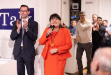 Eine Frau in einem orangefarbenen Anzug spricht in ein Mikrofon über 1 Million Euro für Landesstraßen Steiermark, während die Menschen um sie herum bei einer Indoor-Veranstaltung applaudieren.