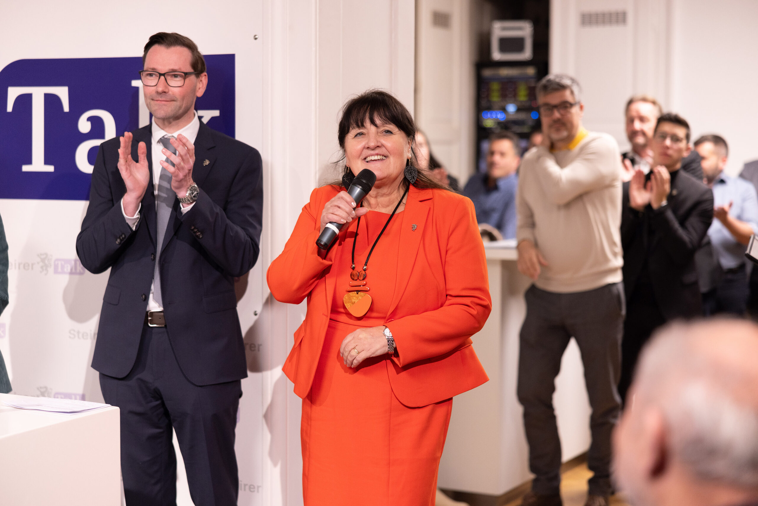 Eine Frau in einem orangefarbenen Anzug spricht in ein Mikrofon über 1 Million Euro für Landesstraßen Steiermark, während die Menschen um sie herum bei einer Indoor-Veranstaltung applaudieren.
