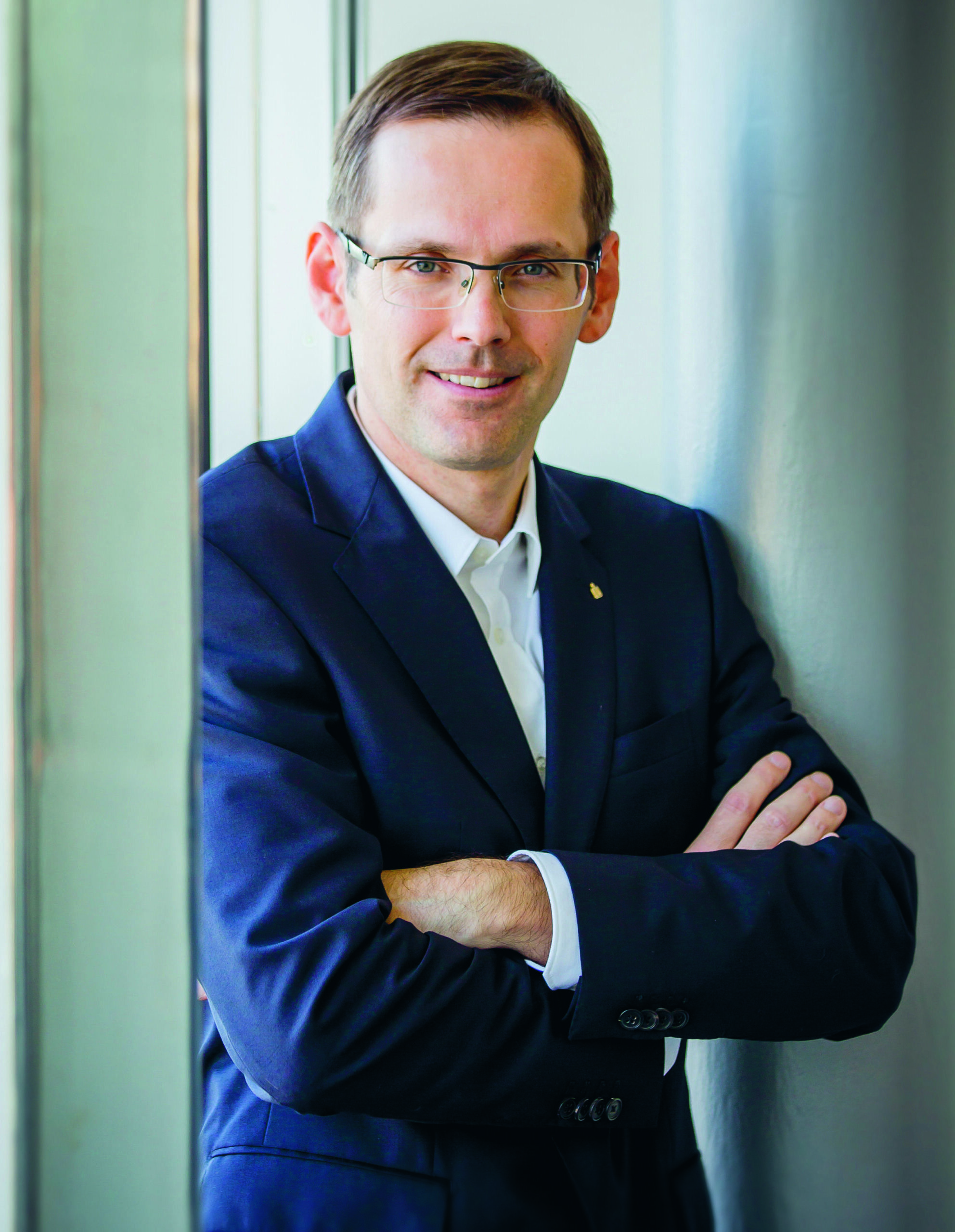 Ein Mann in einem marineblauen Anzug und mit Brille steht mit verschränkten Armen lächelnd neben den großen Fenstern eines hellen, modernen Gebäudes.