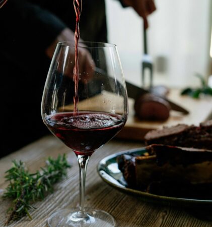 Rotwein, der auf einem Holztisch in ein Glas gegossen wird, während im Hintergrund eine Person Fleisch schneidet und Kräuter in der Nähe sind.