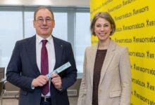 Zwei Personen in Geschäftskleidung lächeln im Haus; der Mann hält eine Karte. Hinter ihnen steht auf einer gelben Wand: Klare Mehrheit für soziale Sicherheit in der Steiermark.