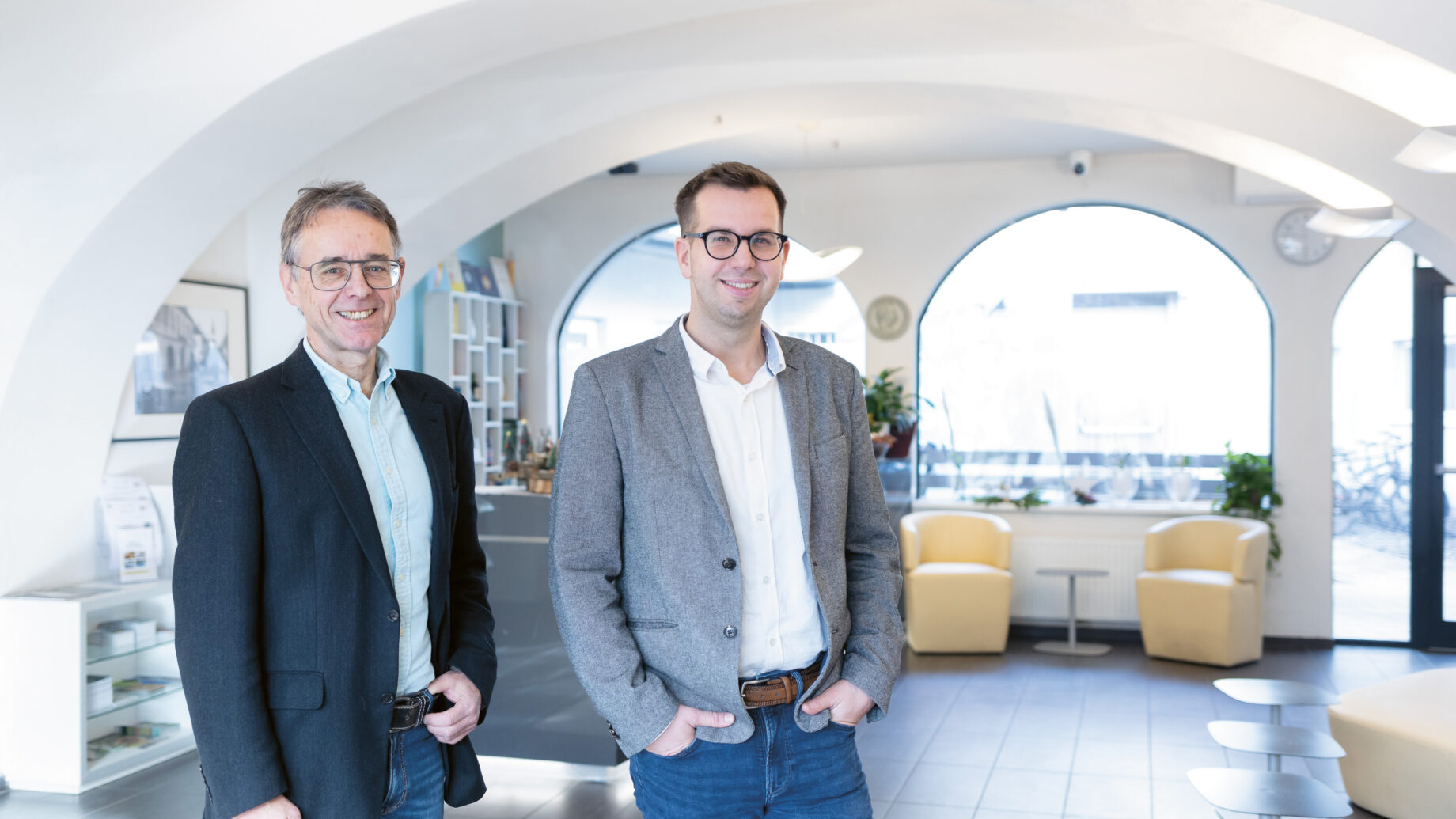 Zwei Männer mit Blazern und Brillen stehen lächelnd in einer hellen, modernen Bürohalle mit Bogenfenstern und gelben Stühlen.