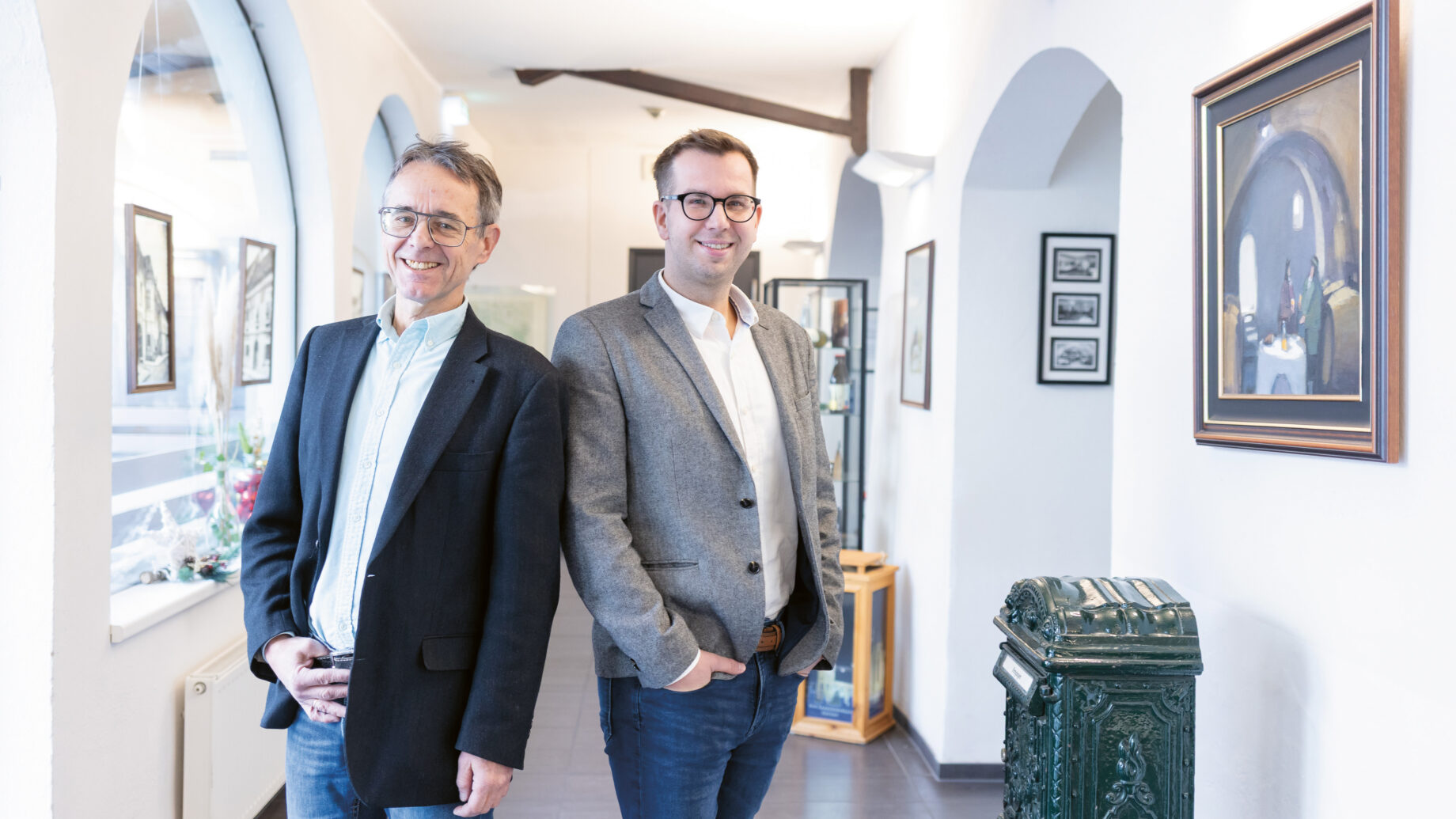 Zwei Männer in Blazern und mit Brille stehen lächelnd in einem hellen Flur mit gewölbten Türen, gerahmten Kunstwerken und Vintage-Dekor.