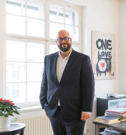 Ein Mann im Anzug steht lächelnd in einem hellen, modernen Büro mit großen Fenstern, einem Schreibtisch, Computerbildschirmen und Kunstwerken an den Wänden.