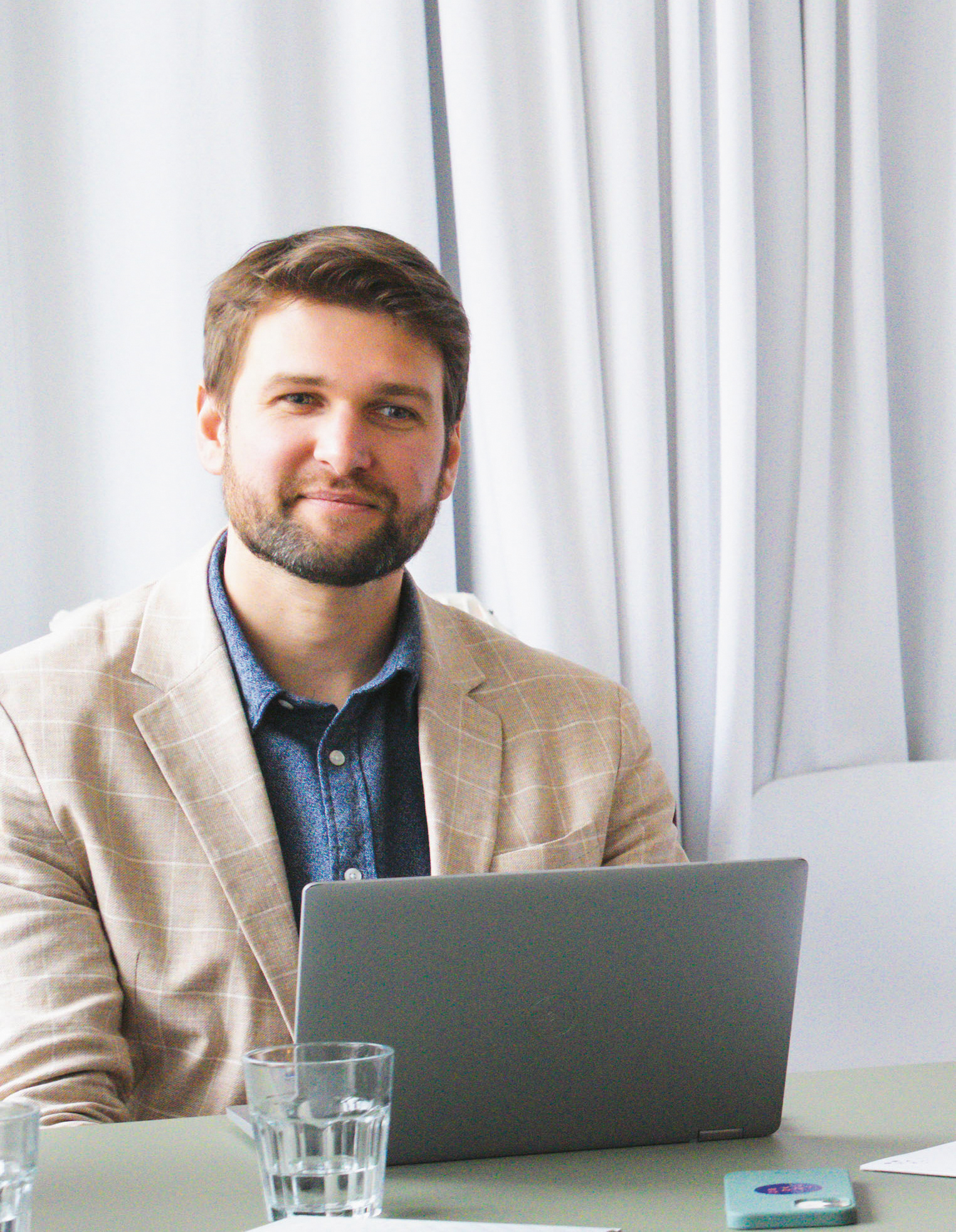 Ein bärtiger Mann im karierten Blazer sitzt an einem Tisch mit einem Laptop, einem Glas Wasser und einer Karte vor einem weißen Vorhang.