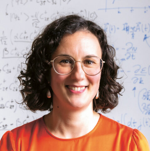 Eine lächelnde Frau mit kurzen lockigen Haaren und Brille, die ein orangefarbenes Oberteil trägt, steht vor einer Tafel mit mathematischen Gleichungen.