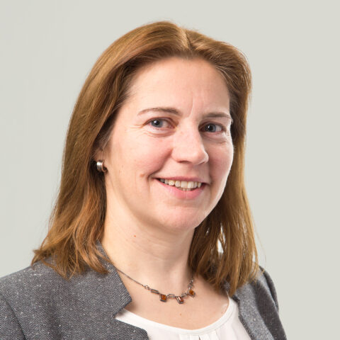 Frau mit schulterlangen braunen Haaren, die einen grauen Blazer, eine weiße Bluse und eine Halskette trägt und in die Kamera lächelt, vor einem schlichten Hintergrund.