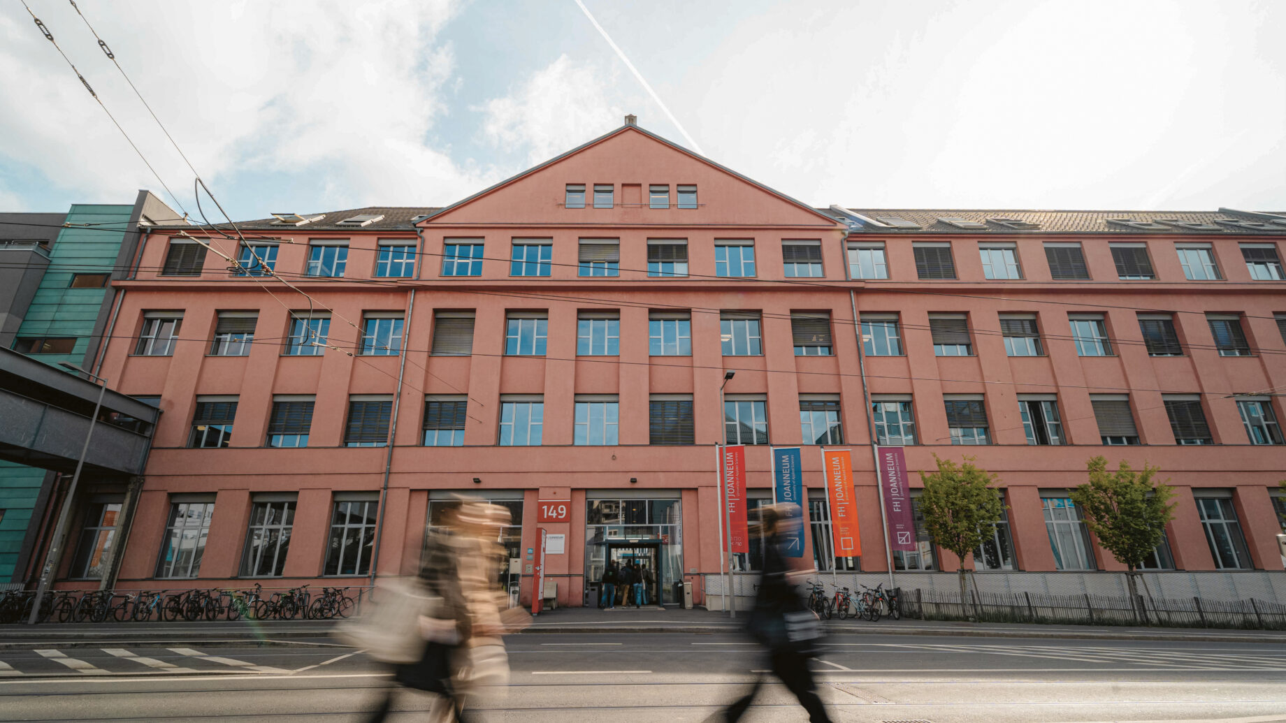 An einem bewölkten Tag gehen Menschen an einem großen rosafarbenen Gebäude mit vielen Fenstern und bunten Bannern vorbei.