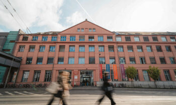 An einem bewölkten Tag gehen Menschen an einem großen rosafarbenen Gebäude mit vielen Fenstern und bunten Bannern vorbei.