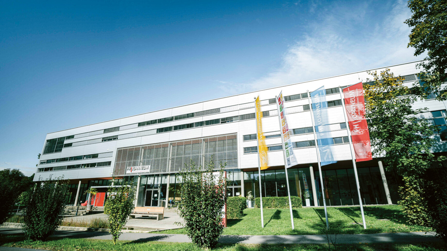 Modernes weißes Gebäude mit farbenfrohen Bannern, großen Fenstern und Grünpflanzen vor dem Gebäude unter einem klaren blauen Himmel.