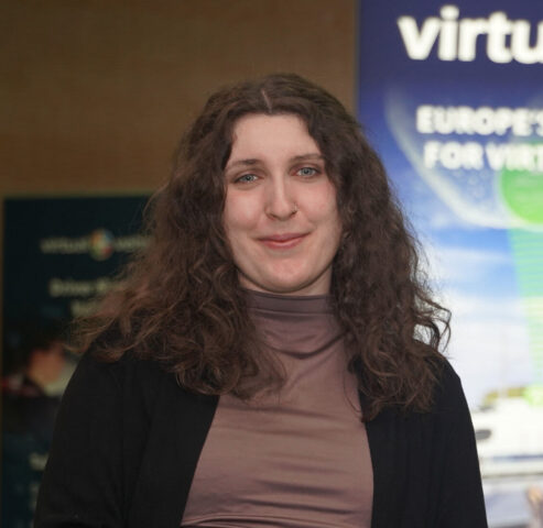 Frau, die an einem Tisch steht und ein kleines Gerät hält, mit einer Pflanze und einem Banner des Forschungszentrums "Virtual Vehicle" im Hintergrund.