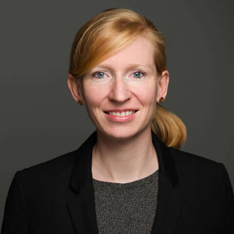 Eine Frau mit blondem Haar in einem Pferdeschwanz, die einen schwarzen Blazer und ein gemustertes Oberteil trägt, lächelt in die Kamera vor einem schlichten grauen Hintergrund.