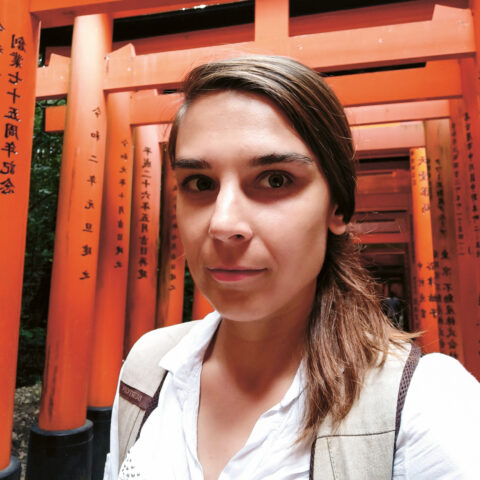 Eine Frau mit braunen Haaren macht ein Selfie vor einem orangefarbenen Torii-Tor mit japanischer Schrift am Fushimi-Inari-Schrein in Kyoto, Japan.