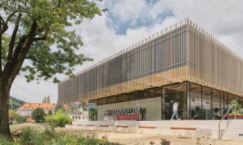 Modernes Gebäude mit vertikalen Metalllamellen, großen Glasfenstern und einem Schild mit der Aufschrift "FH JOANNEUM University of Applied Sciences"; Person im Freien.