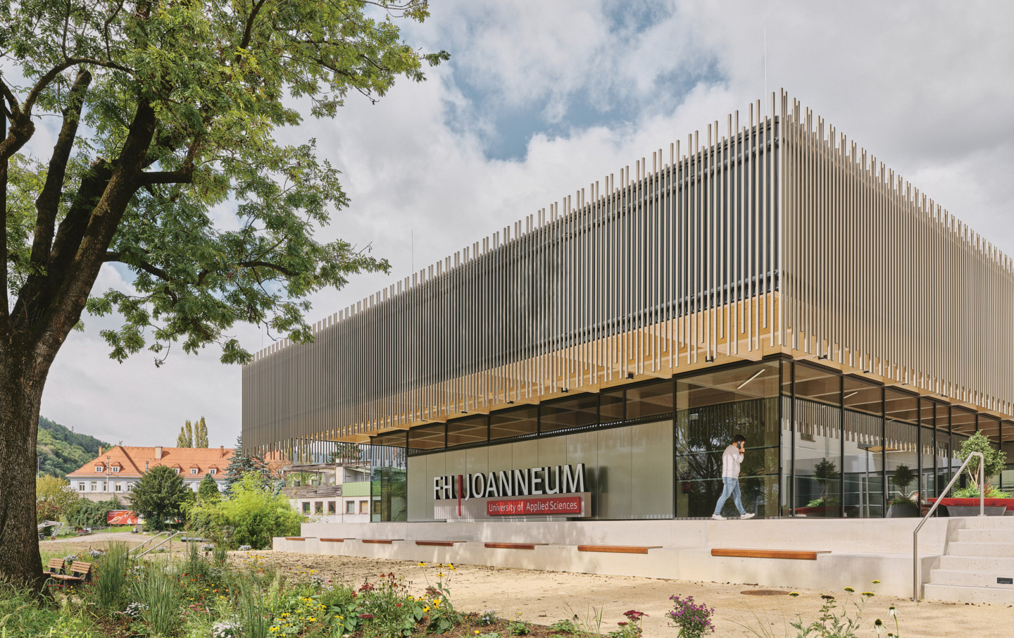 Modernes Gebäude mit vertikalen Metalllamellen, großen Glasfenstern und einem Schild mit der Aufschrift "FH JOANNEUM University of Applied Sciences"; Person im Freien.