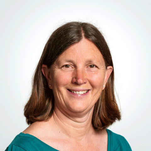 Eine Frau mittleren Alters mit glattem braunem Haar lächelt in die Kamera und trägt ein blaugrünes Oberteil vor einem schlichten hellen Hintergrund.