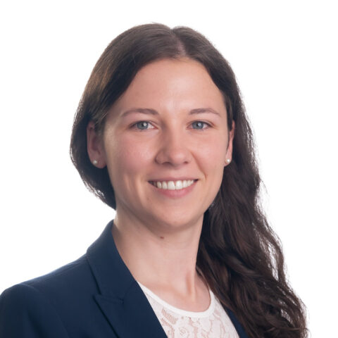 Eine Frau mit langen braunen Haaren trägt einen marineblauen Blazer und eine weiße Bluse und lächelt in die Kamera vor einem schlichten weißen Hintergrund.