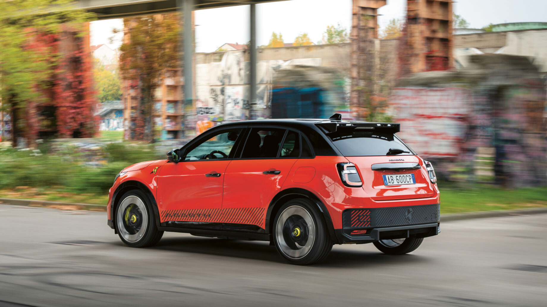 Ein rot-schwarzer Geländewagen fährt auf einer Stadtstraße mit unscharfen, mit Graffiti beschmierten Wänden und Grünflächen im Hintergrund.