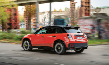 Ein rot-schwarzer Geländewagen fährt auf einer Stadtstraße mit unscharfen, mit Graffiti beschmierten Wänden und Grünflächen im Hintergrund.