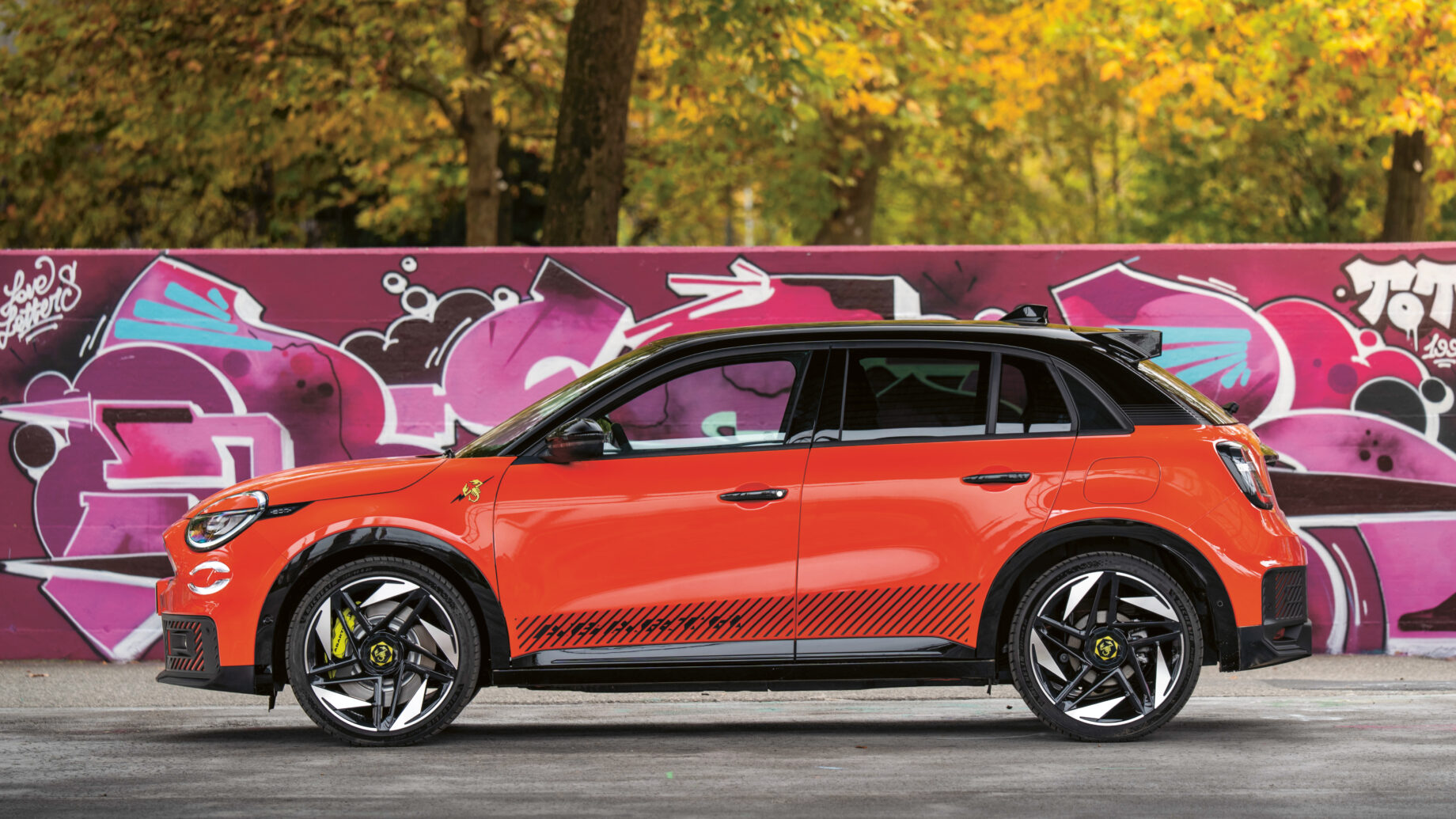 Ein leuchtend orangefarbener Geländewagen parkt vor einer lilafarbenen Graffitiwand mit Bäumen mit Herbstlaub im Hintergrund.