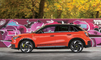 Ein leuchtend orangefarbener Geländewagen parkt vor einer lilafarbenen Graffitiwand mit Bäumen mit Herbstlaub im Hintergrund.