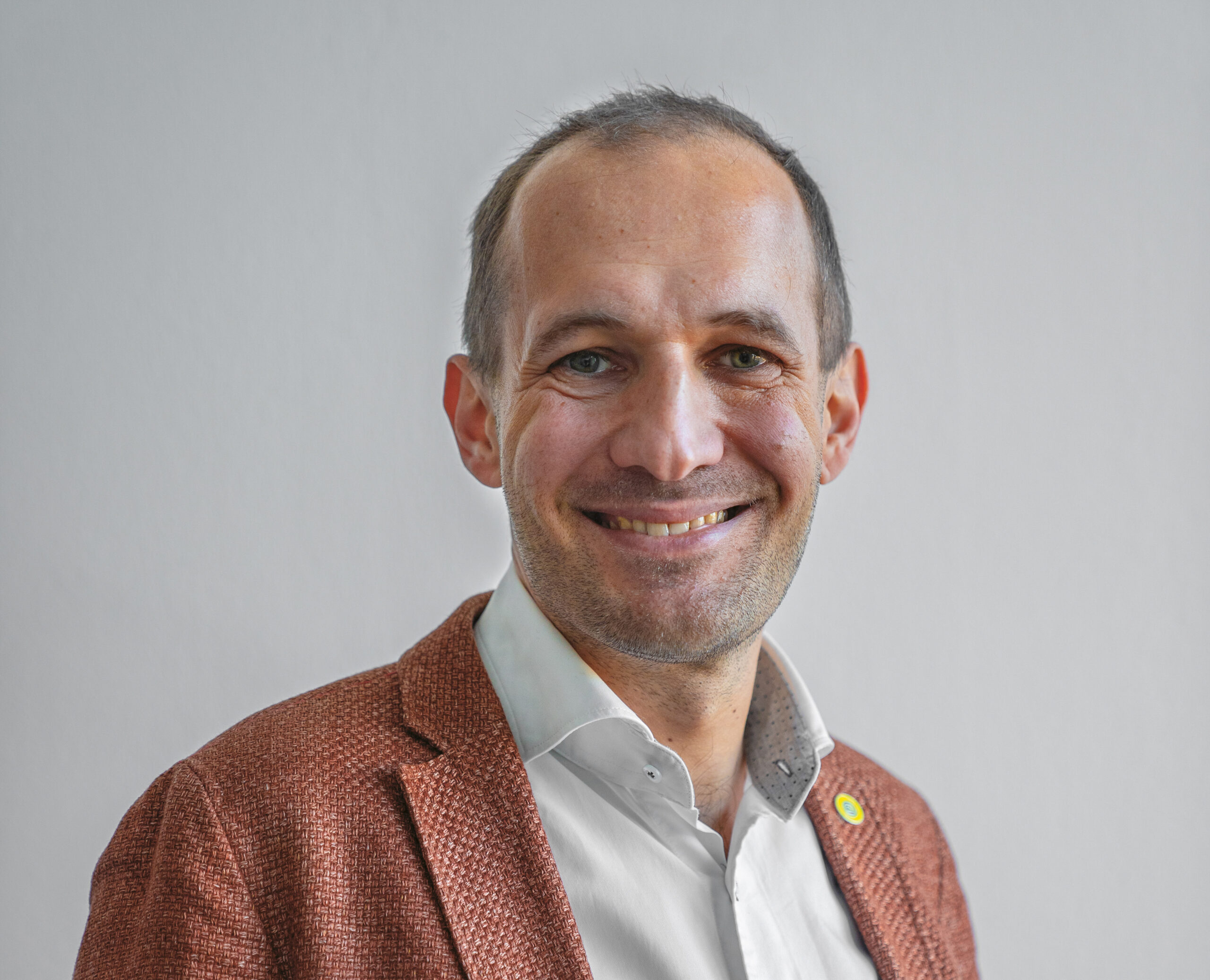 Ein Mann mit kurzen Haaren lächelt, trägt ein weißes Hemd und einen strukturierten braunen Blazer und steht vor einem schlichten hellgrauen Hintergrund.