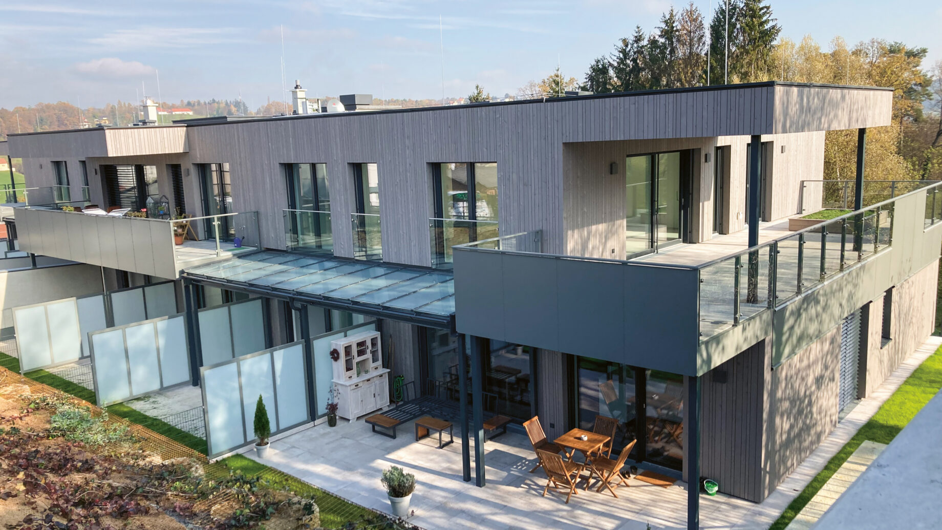Modernes zweistöckiges Doppelhaus mit großen Balkonen, Glasgeländern und einer geräumigen Terrasse mit Gartenmöbeln, umgeben von Grünflächen und Gartenbeeten.