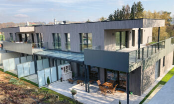 Modernes zweistöckiges Doppelhaus mit großen Balkonen, Glasgeländern und einer geräumigen Terrasse mit Gartenmöbeln, umgeben von Grünflächen und Gartenbeeten.