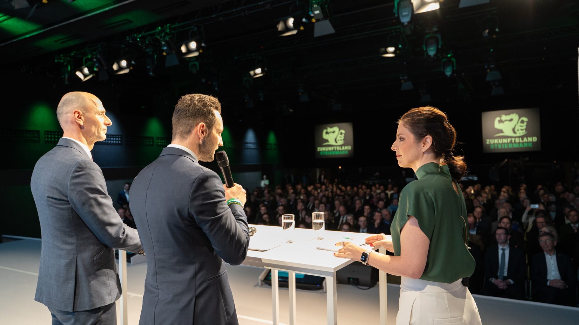 Drei Personen in Business-Kleidung sprechen auf einer Konferenz über "Die klare Mehrheit für soziale Sicherheit in der Steiermark" vor einem großen Publikum.