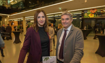 Eine Frau in einem lilafarbenen Blazer und ein Mann in einem grauen Anzug stehen bei einer Indoor-Veranstaltung mit Menschen und Tischen im Hintergrund zusammen.