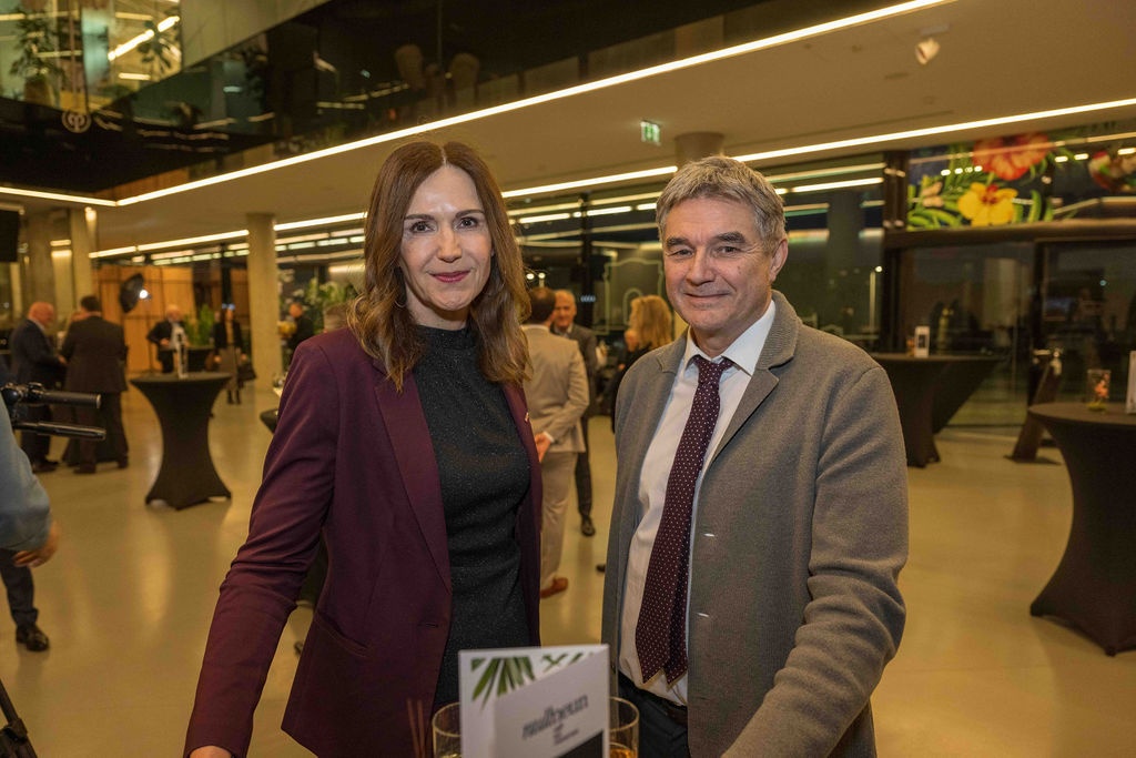 Eine Frau in einem lilafarbenen Blazer und ein Mann in einem grauen Anzug stehen bei einer Indoor-Veranstaltung mit Menschen und Tischen im Hintergrund zusammen.