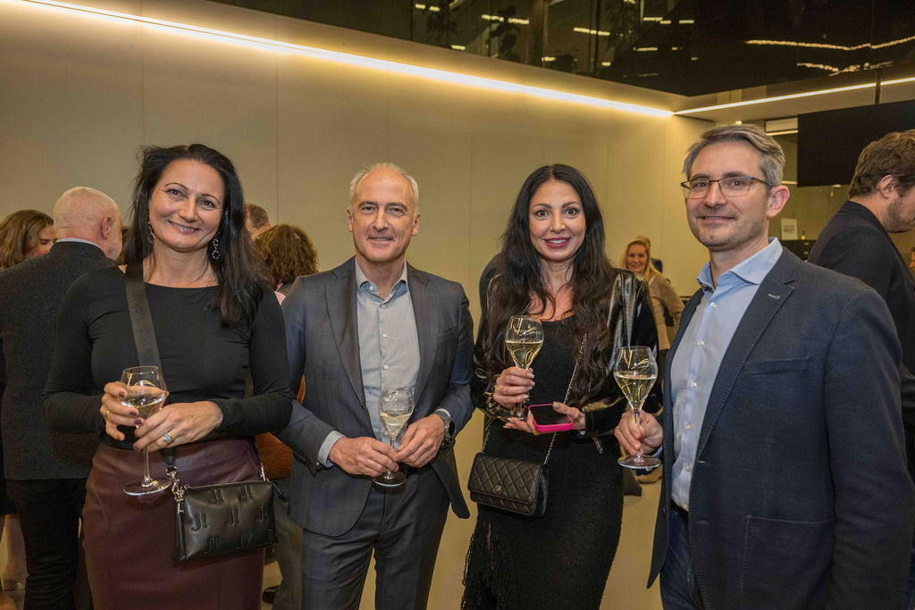 Vier formell gekleidete Personen stehen bei einer Veranstaltung in einer Halle zusammen, lächeln und halten Gläser mit Weißwein in der Hand, während sich andere im Hintergrund unter sie mischen.