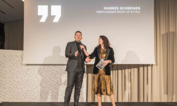 Ein Mann in einem dunklen Anzug und eine Frau in einem goldenen Kleid stehen auf der Bühne und sprechen in Mikrofone vor einem Bildschirm mit der Aufschrift "Hannes Schreiner".
