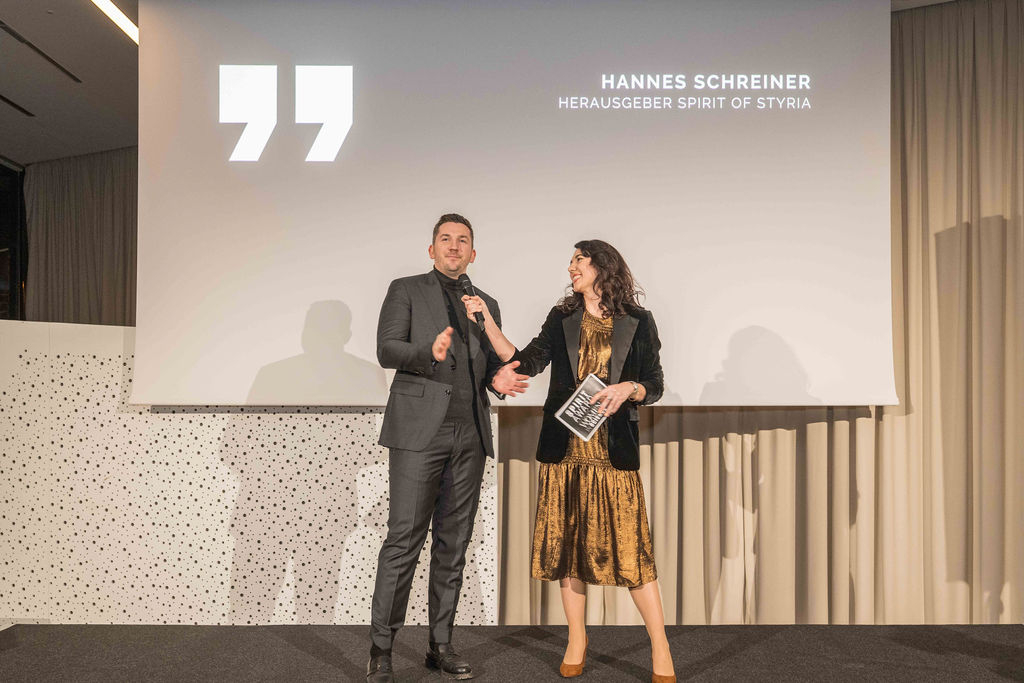 Ein Mann in einem dunklen Anzug und eine Frau in einem goldenen Kleid stehen auf der Bühne und sprechen in Mikrofone vor einem Bildschirm mit der Aufschrift "Hannes Schreiner".