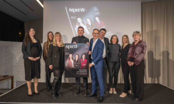 Neun Personen stehen in einem Innenraum zusammen, lächeln und halten ein großes Exemplar eines Magazintitels mit der Überschrift "spirit" in der Hand, dahinter ein schlichter Hintergrund.
