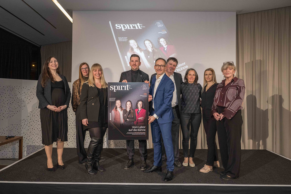 Neun Personen stehen in einem Innenraum zusammen, lächeln und halten ein großes Exemplar eines Magazintitels mit der Überschrift "spirit" in der Hand, dahinter ein schlichter Hintergrund.