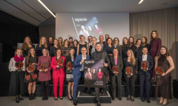 Eine Gruppe von Frauen und zwei Männern posieren mit Trophäen und einem großen Zeitschriftencover auf der Bühne und feiern bei einer Preisverleihung.