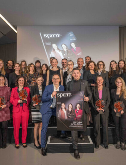 Eine große Gruppe von Menschen, die in einem Raum posieren, viele von ihnen mit Preisen in der Hand, während hinter ihnen auf einem Bildschirm das Titelbild eines Magazins zu sehen ist, das auf einer Preisverleihungsveranstaltung erscheint.