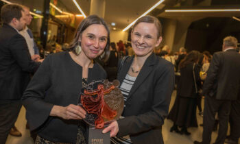 Zwei Frauen lächeln bei einer Veranstaltung, eine hält eine Spirit Award-Trophäe. Im Hintergrund mischen sich Menschen in einem modernen, gut beleuchteten Veranstaltungsort.