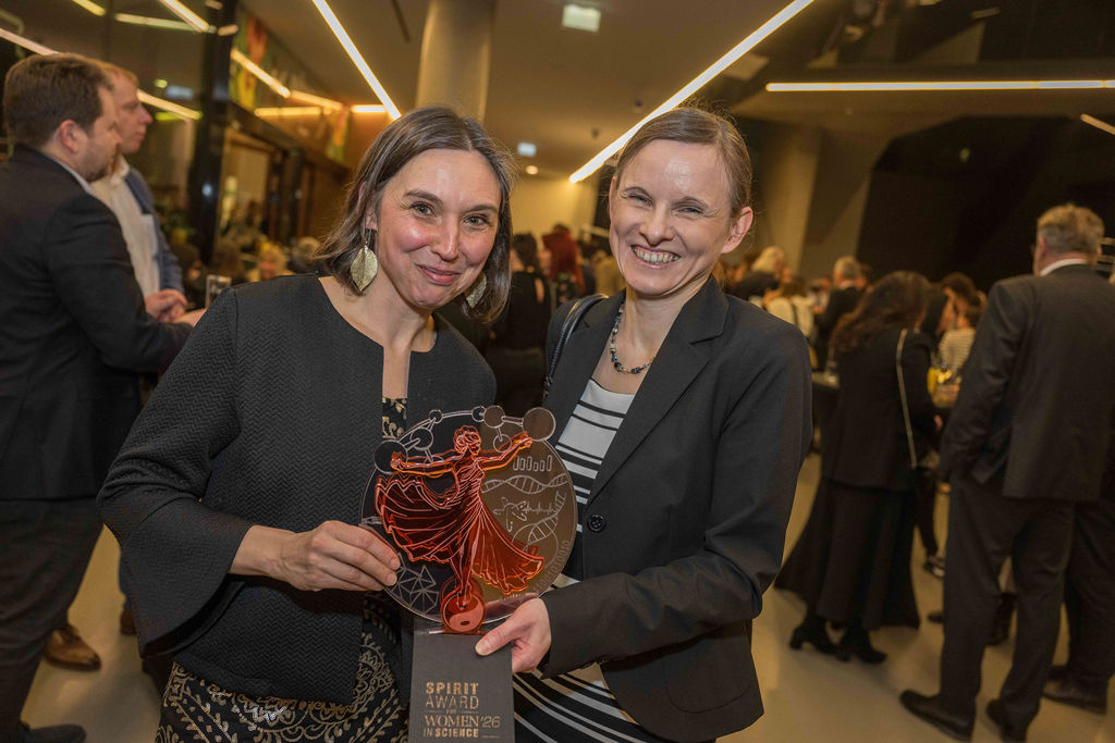 Zwei Frauen lächeln bei einer Veranstaltung, eine hält eine Spirit Award-Trophäe. Im Hintergrund mischen sich Menschen in einem modernen, gut beleuchteten Veranstaltungsort.